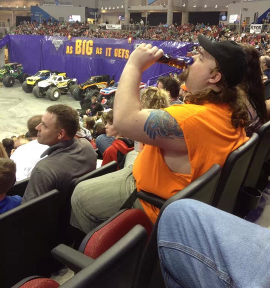 Overweight white male with a mullet and a trucker hat drinking a bottle of syrup at a monster truck rally